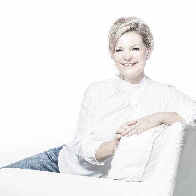 Silvia Ricanek Frau in weißem Hemd sitzt entspannt auf einer hellen Couch und lächelt in die Kamera.